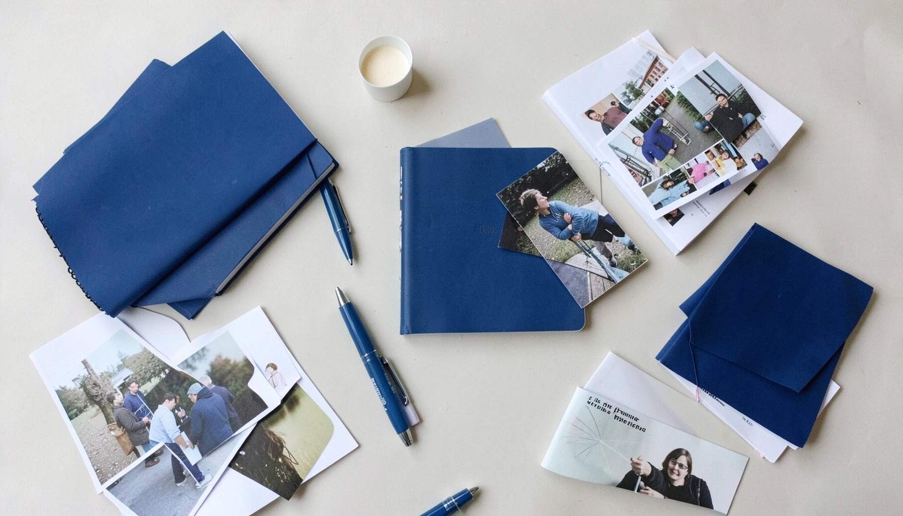Overhead flat-lay photograph of notebooks, pens, and printed photographs spread across a light surface, documenting community work.