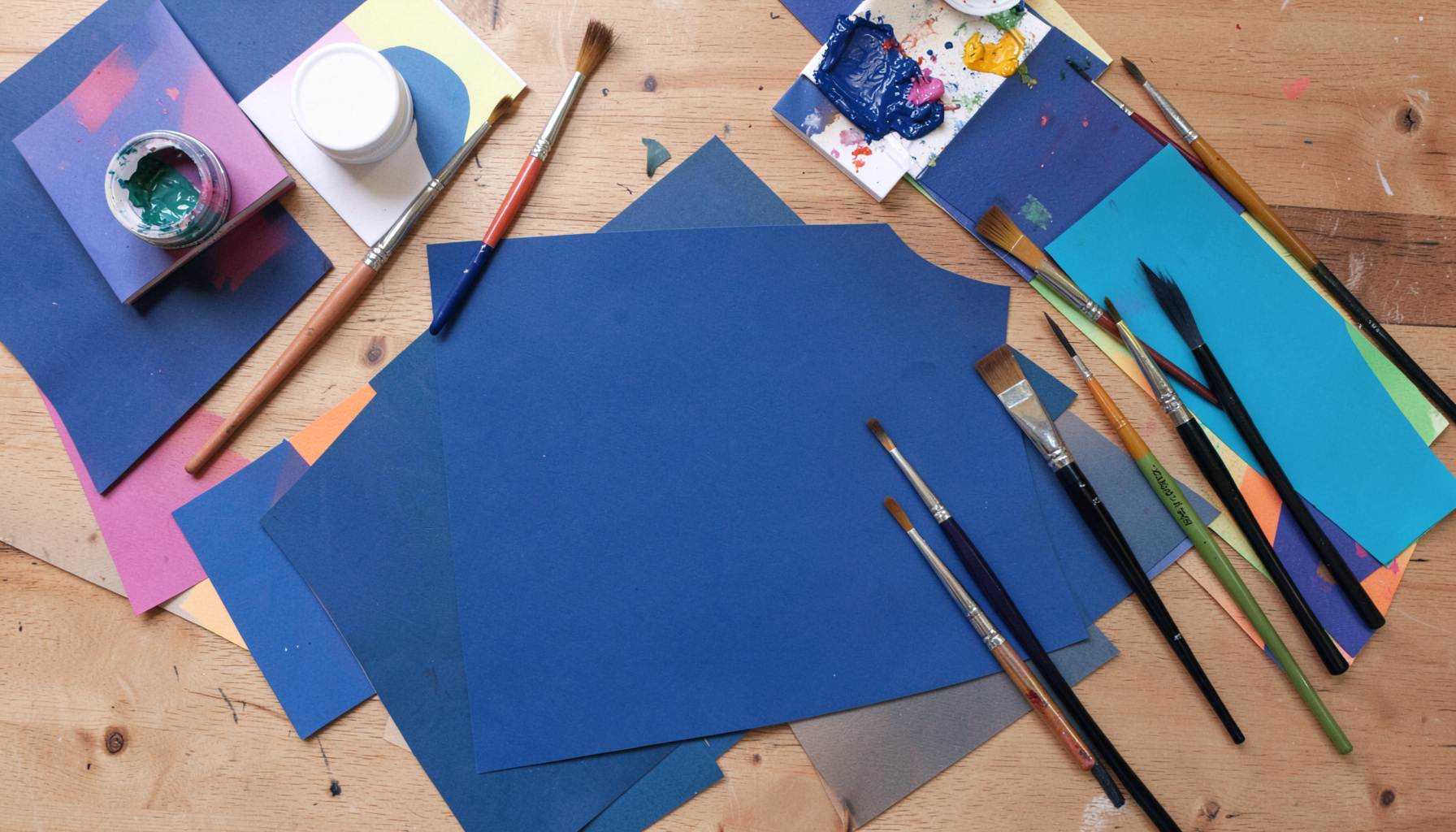 Overhead flat-lay photograph of art supplies, paintbrushes, and colorful paper arranged on a warm wooden table, creative mood
