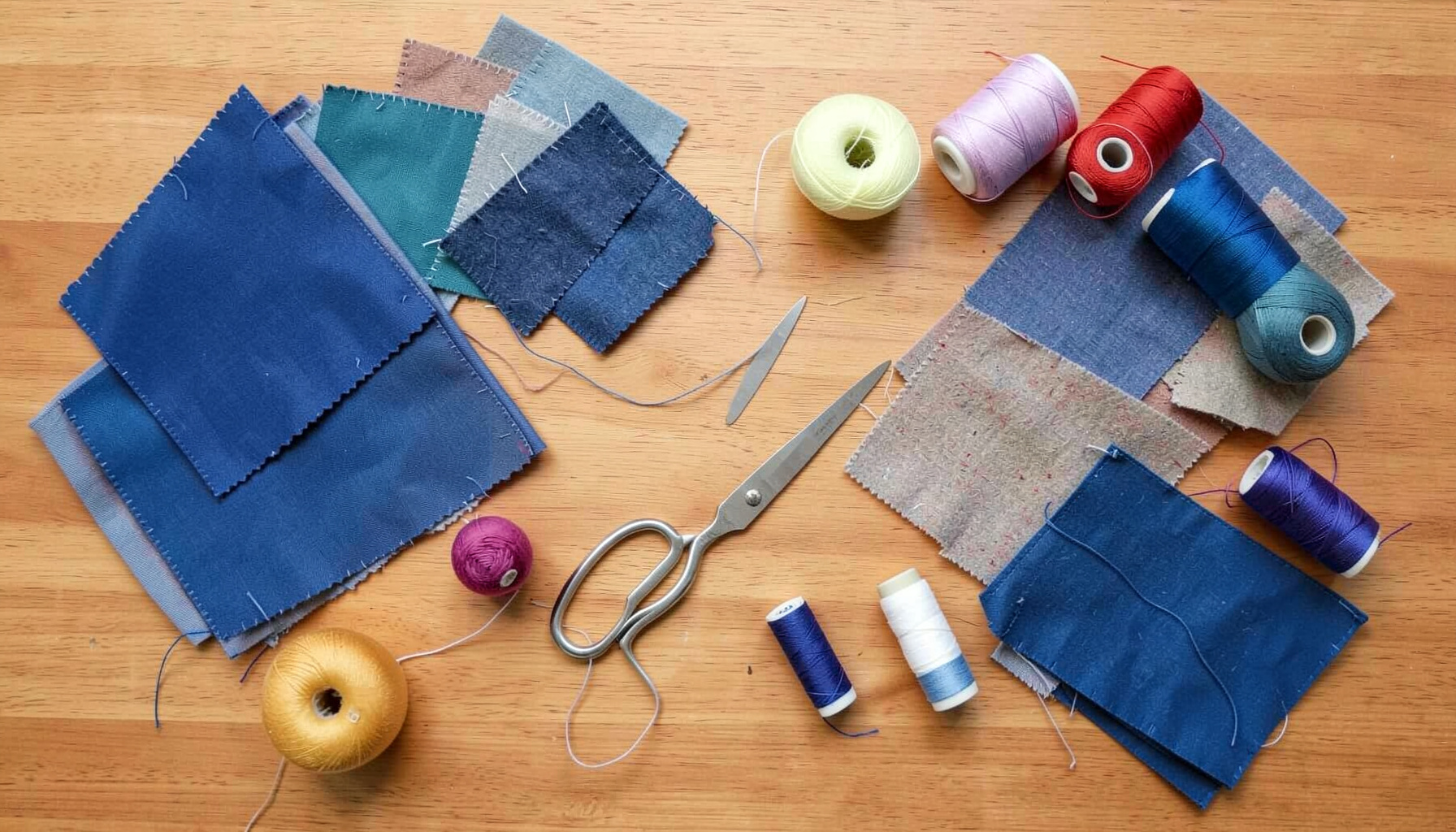 Overhead flat-lay photograph of vibrant craft supplies, fabric swatches, scissors, and colorful thread spools arranged on a wooden surface.
