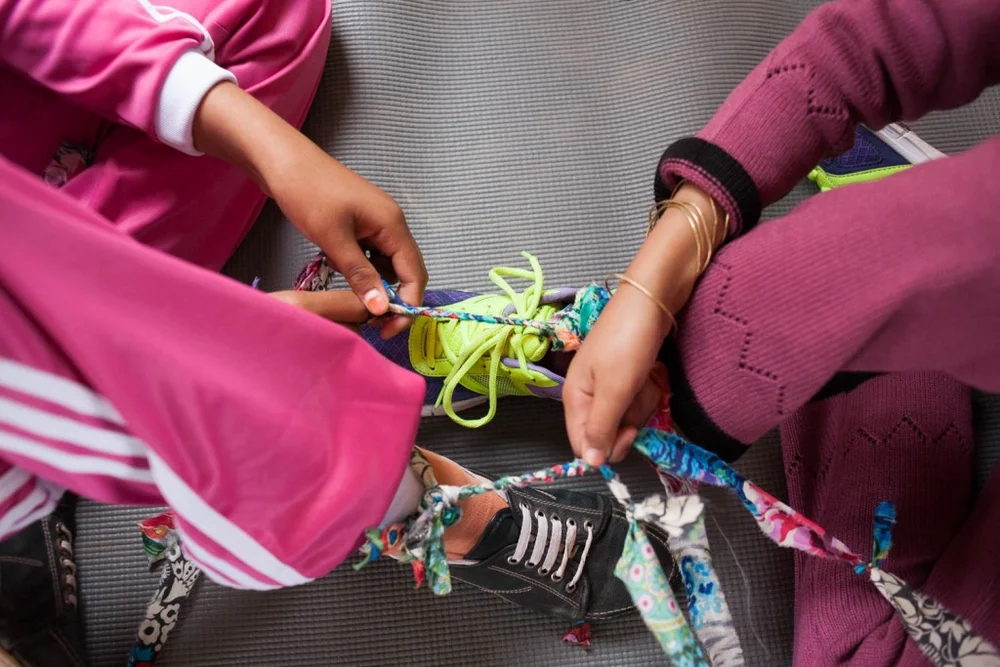 Close-up overhead shot of hands tying colorful fabric strips and shoelaces together, pink and teal tones, playful cooperative mood