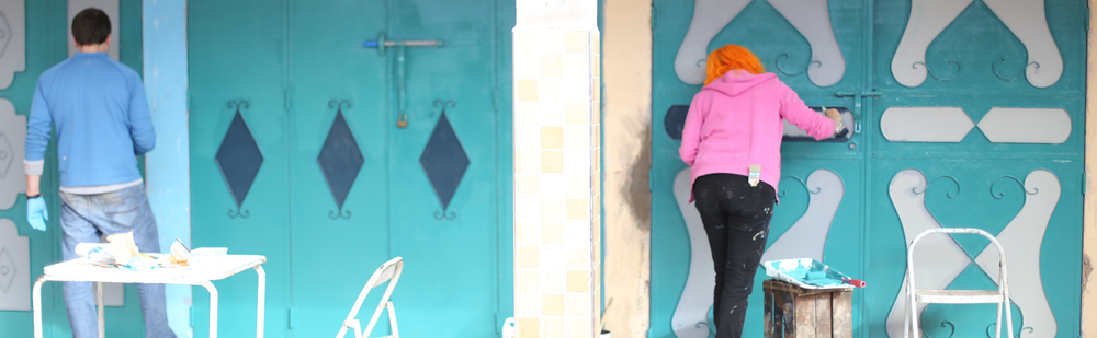 Two people painting ornate teal doors with white decorative patterns, wearing pink and blue clothing, a busy and creative scene